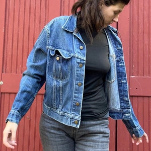 May include: A woman wearing a blue denim jacket over a black mock neck shirt and grey jeans. The jacket has a button closure and a pocket on the left chest. The woman is standing in front of a red wooden door.