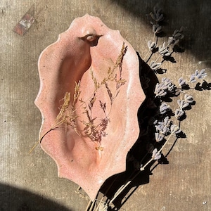 May include: A handmade, pink ceramic dish with an organic, scalloped edge. The dish is decorated with dried grasses and lavender sprigs. The dish is displayed on a weathered wooden surface, with shadows cast across the scene.