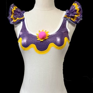 May include: A white tank top with purple and yellow trim. The top has a yellow and white sun design with a pink pom-pom in the center.
