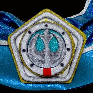 May include: A gold star-shaped ornament with a silver Rebel Alliance symbol in the center. The symbol is surrounded by a white circle with a red stripe at the bottom. The ornament is covered in glitter.