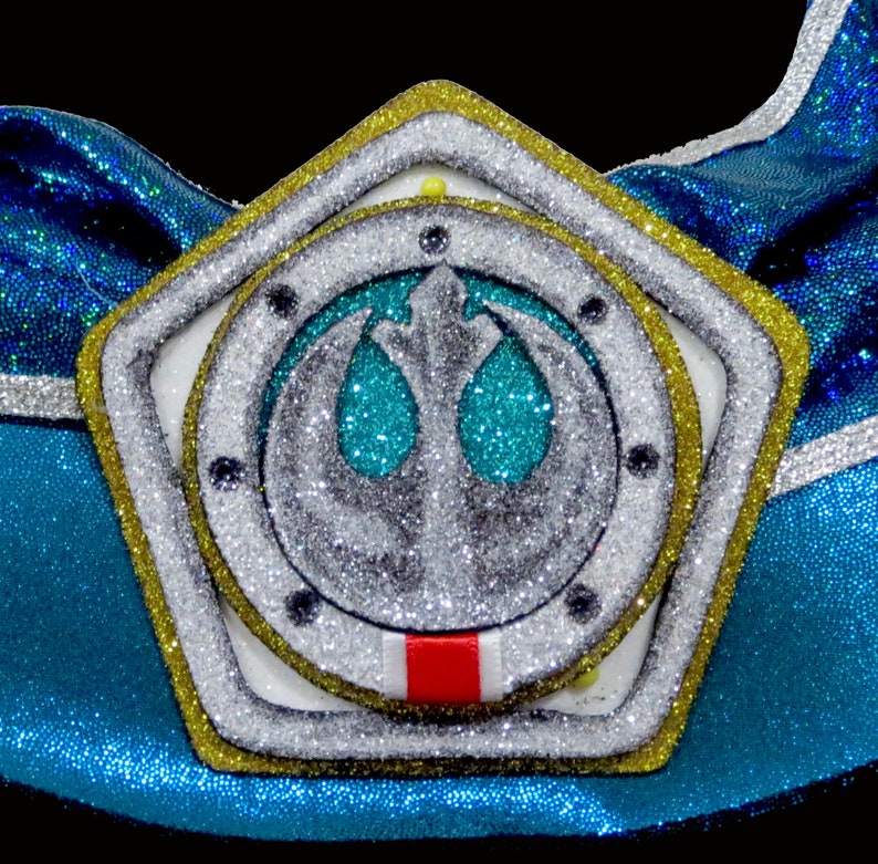 May include: A gold star-shaped ornament with a silver Rebel Alliance symbol in the center. The symbol is surrounded by a white circle with a red stripe at the bottom. The ornament is covered in glitter.