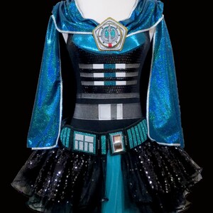 May include: A teal and black sequined costume with a cape and a tutu skirt. The costume features a silver and black design on the bodice and a silver emblem with a symbol on the chest.
