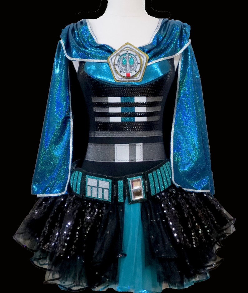 May include: A teal and black sequined costume with a cape and a tutu skirt. The costume features a silver and black design on the bodice and a silver emblem with a symbol on the chest.