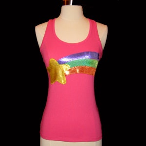 May include: A pink tank top with a gold star and a rainbow design. The star is on the left side, and the rainbow stretches across the chest. The tank top is displayed on a mannequin.