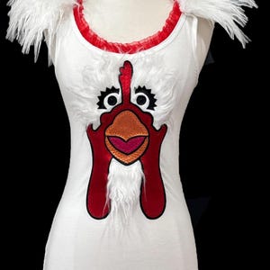 May include: White tank top with a red and orange rooster face graphic. The rooster has white fur around its neck and white feathers on the shoulders.