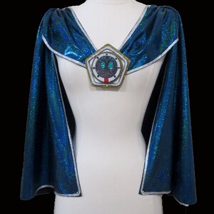 May include: A blue cape with silver trim and a large, round, silver and gold brooch with a green and black design in the center.