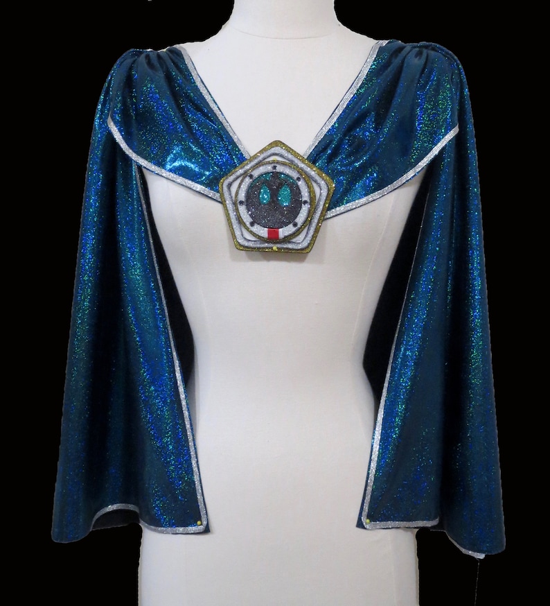 May include: A blue cape with silver trim and a large, round, silver and gold brooch with a green and black design in the center.