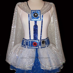May include: A white and blue glittery costume with a white tulle skirt, a cape, and a belt. The costume is decorated with silver and blue glitter and features a blue and silver design on the bodice. The costume is inspired by the character R2-D2 from the Star Wars franchise.