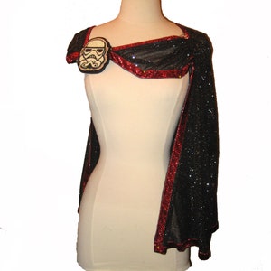 May include: A black and red cape with a sparkly texture. The cape has a red trim and a white and black Stormtrooper helmet patch on the shoulder.