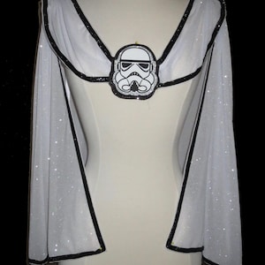 May include: White cape with black trim and a Stormtrooper emblem. The cape is made of a sheer, sparkly fabric and is designed to be worn over the shoulders. The emblem is a black and white graphic of a Stormtrooper helmet.