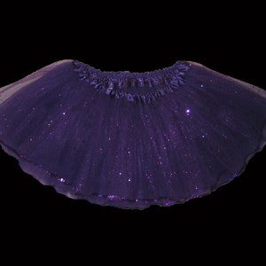 May include: A dark purple tutu skirt with a shimmering, glittery effect. The skirt has a ruffled waistband and a scalloped hemline. The tulle fabric is layered, creating a full, flowing silhouette. Perfect for dance or dress-up.