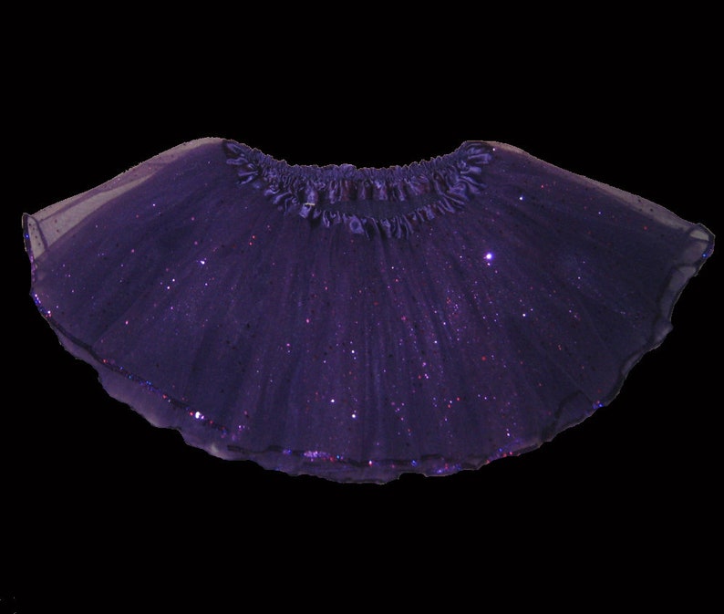 May include: A dark purple tutu skirt with a shimmering, glittery effect. The skirt has a ruffled waistband and a scalloped hemline. The tulle fabric is layered, creating a full, flowing silhouette. Perfect for dance or dress-up.
