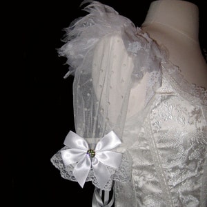 May include: White wedding dress with a lace bodice and tulle sleeves. The sleeves have a white satin bow with a rhinestone embellishment.
