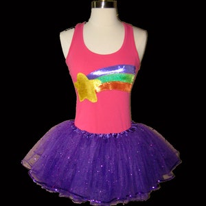May include: A pink tank top with a gold star and rainbow sequin design, paired with a purple tutu skirt. The costume is displayed on a mannequin. The tutu is made of layers of purple tulle with glitter.