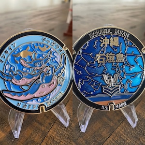 May include: Two-sided coin with a black border. One side features a blue and pink manta ray design with Japanese text. The other side has a blue constellation design with Japanese text and an eagle emblem. The coin is displayed on a clear stand.