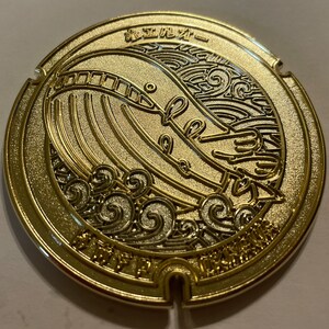 May include: A gold-colored, circular metal object with a detailed relief design. The design features a whale, waves, and Japanese text. The object has a decorative, ornate appearance, with a shiny, reflective surface.