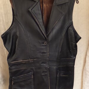May include: Black leather vest with a notched collar, button closure, and two front pockets. The vest has a brown interior lining and is hanging on a metal hanger.