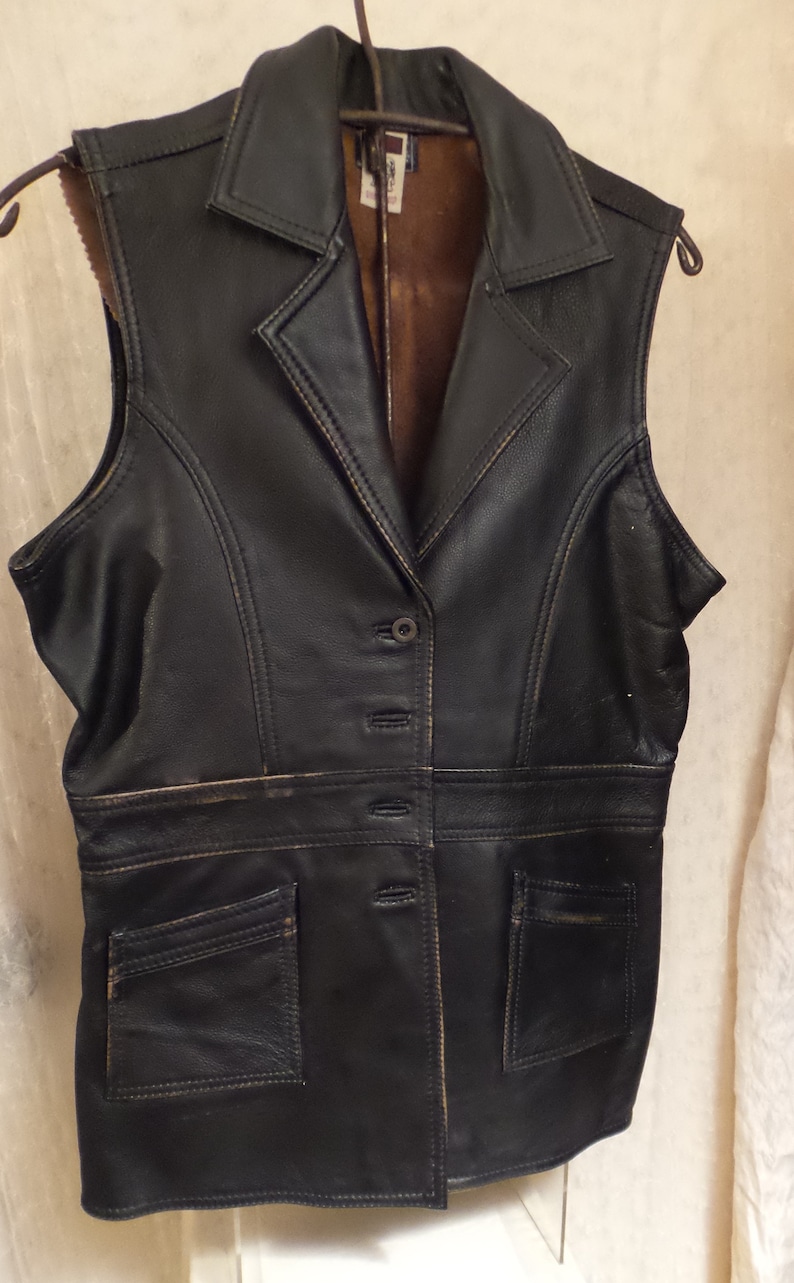 May include: Black leather vest with a notched collar, button closure, and two front pockets. The vest has a brown interior lining and is hanging on a metal hanger.