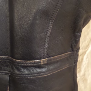 May include: Close-up of a section of a black leather jacket. The jacket features visible stitching and a horizontal seam across the midsection. The leather has a slightly distressed appearance with some lighter areas.