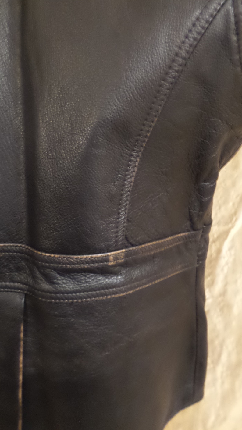 May include: Close-up of a section of a black leather jacket. The jacket features visible stitching and a horizontal seam across the midsection. The leather has a slightly distressed appearance with some lighter areas.