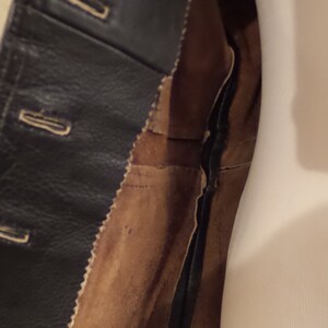 May include: Close-up of a black leather jacket with button closures and a brown suede lining. The jacket's interior is visible, showcasing the stitching and construction details. The garment is likely vintage or well-worn.