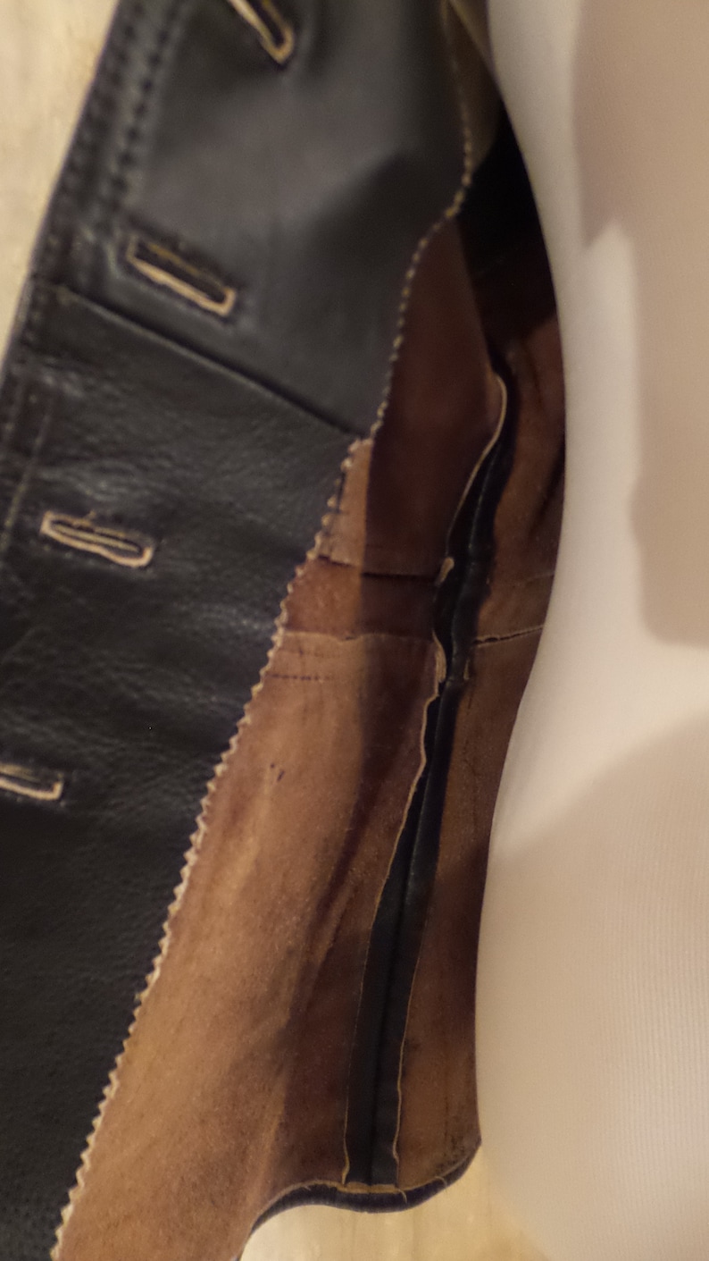 May include: Close-up of a black leather jacket with button closures and a brown suede lining. The jacket's interior is visible, showcasing the stitching and construction details. The garment is likely vintage or well-worn.