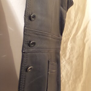 May include: Black leather vest with a tailored fit. The vest features a button-up front, a small pocket, and decorative stitching along the seams. The vest is sleeveless and appears to be made of genuine leather.
