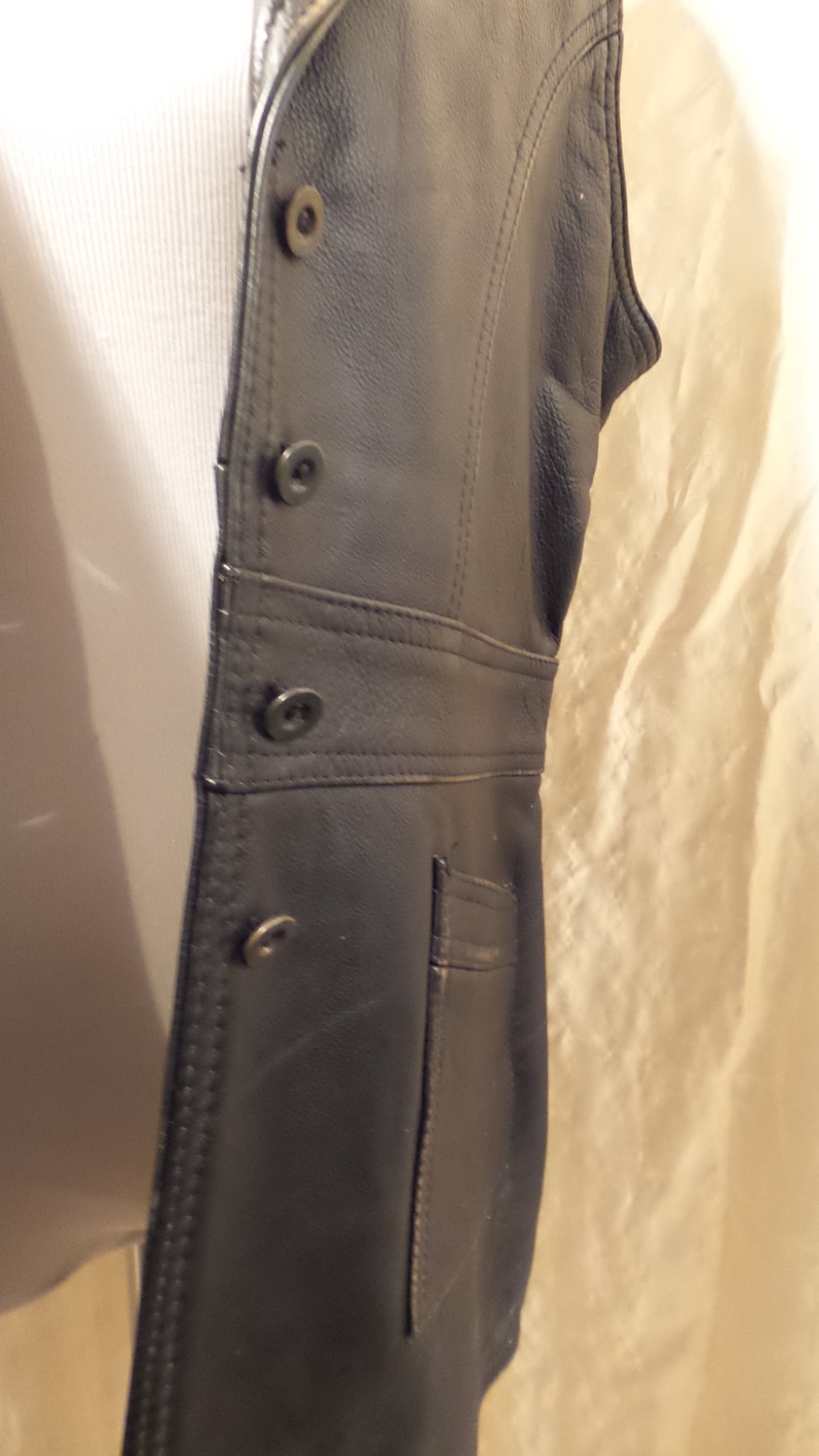 May include: Black leather vest with a tailored fit. The vest features a button-up front, a small pocket, and decorative stitching along the seams. The vest is sleeveless and appears to be made of genuine leather.
