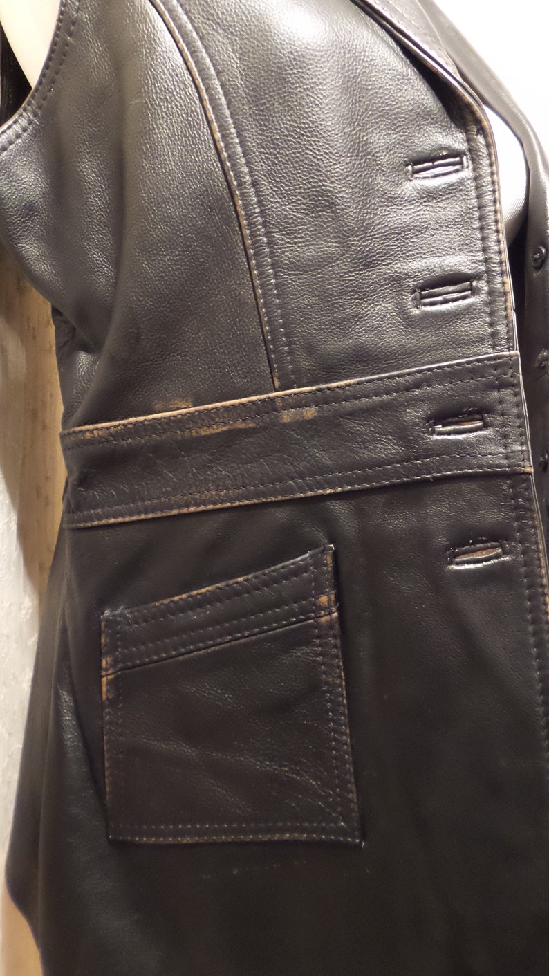 May include: Black leather vest with a tailored fit. Features include a notched lapel, button closure, and a single pocket. The vest has visible stitching and a belt detail at the waist. The leather shows signs of wear.