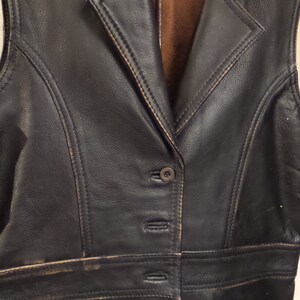May include: A black leather vest with a notched collar and a button-down front. The vest has two front pockets and a brown interior lining. The leather shows signs of wear, with distressed edges and subtle color variations.