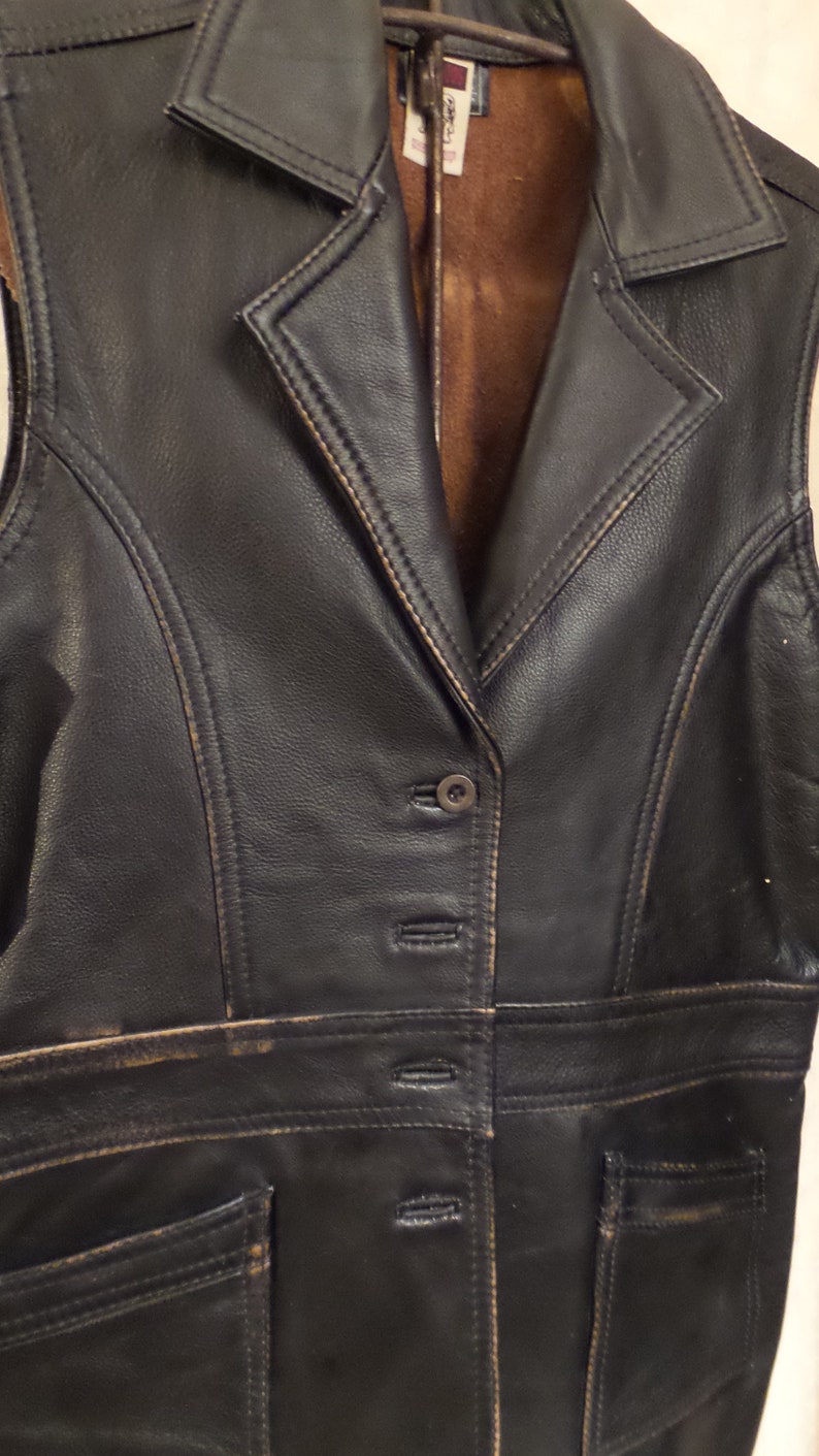 May include: A black leather vest with a notched collar and a button-down front. The vest has two front pockets and a brown interior lining. The leather shows signs of wear, with distressed edges and subtle color variations.