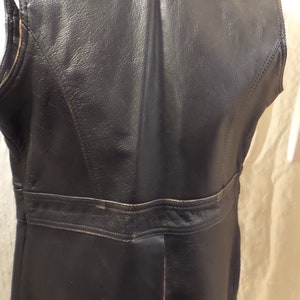 May include: A black leather vest with a collar and a tailored fit. The vest has a classic design with visible stitching and a belt detail at the waist. The leather appears to be slightly worn, adding character to the garment.