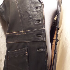 May include: A black leather vest with a notched collar, button closure, and a small pocket. The vest has a tailored fit and a distressed, vintage appearance. The stitching is visible, and the edges show wear.