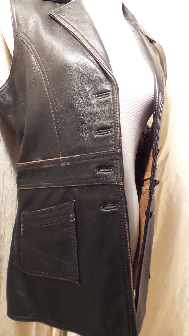 May include: A black leather vest with a notched collar, button closure, and a small pocket. The vest has a tailored fit and a distressed, vintage appearance. The stitching is visible, and the edges show wear.