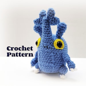 May include: A blue crocheted toy creature with large black eyes, yellow eye accents, and white feet. The text "Crochet Pattern" is displayed in the lower left corner. The handmade toy is on a white background.
