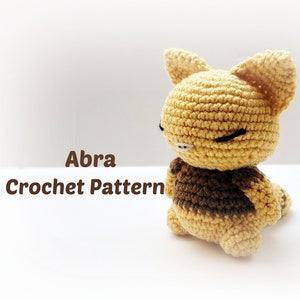Könnte beinhalten: Ein gehäkeltes Plüschtier in Form einer braunen und gelben Katze. Das Spielzeug sitzt auf einer weißen Oberfläche. Der Text "Abra Crochet Pattern" ist unter dem Spielzeug sichtbar.