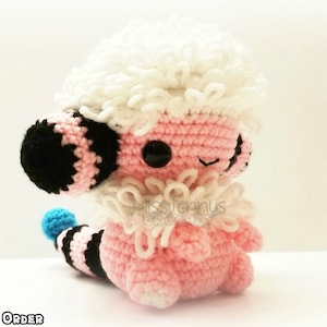 May include: A handmade crochet plush toy in the shape of a cartoon character. The toy is primarily pink with a white, fluffy head and collar. It has black and white striped details, black eyes, and a blue tail tip. The text "MADE TO ORDER" is at the bottom.