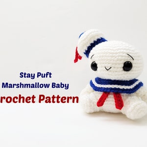May include: A white crocheted Stay Puft Marshmallow Man baby doll with a blue and white sailor collar and red bow tie. The doll has black eyes and a smiling face. The text "Stay Puft Marshmallow Baby Crochet Pattern" is written below the doll.