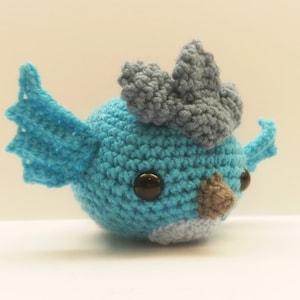 May include: A handmade crochet bird in shades of blue, gray, and brown. The bird has large black eyes and a small beak. The wings and head feathers are detailed. The text "MADE TO ORDER" is at the bottom.
