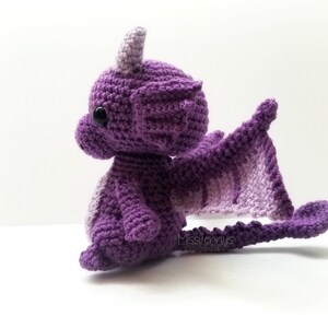 May include: A handmade crochet dragon in shades of purple. The dragon has large wings, a long tail, and small horns. The dragon is sitting on a white surface. The text "MissJenny's Crochet" is visible on the image.