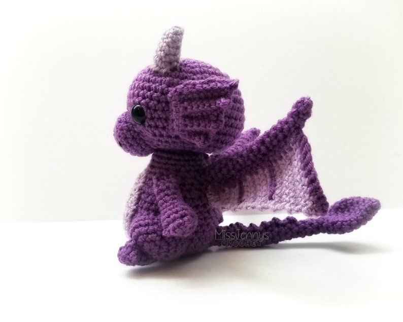 May include: A handmade crochet dragon in shades of purple. The dragon has large wings, a long tail, and small horns. The dragon is sitting on a white surface. The text "MissJenny's Crochet" is visible on the image.