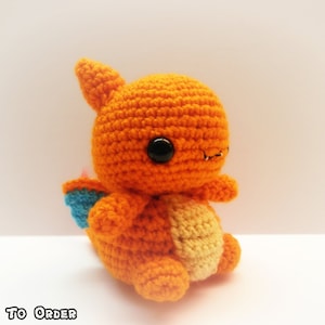 May include: A crocheted orange and yellow Charmander Pokemon plush toy with blue and pink wings. The toy has black eyes and a small, stitched smile.