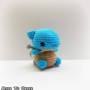 May include: A crocheted blue and brown turtle plush toy with a cute face. The toy is sitting on a white surface.