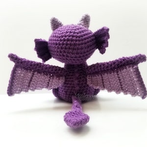 May include: A handmade crochet dragon in shades of purple, viewed from the back. The dragon has outstretched wings, small horns, and a tail. The wings and body are a gradient of purple hues.