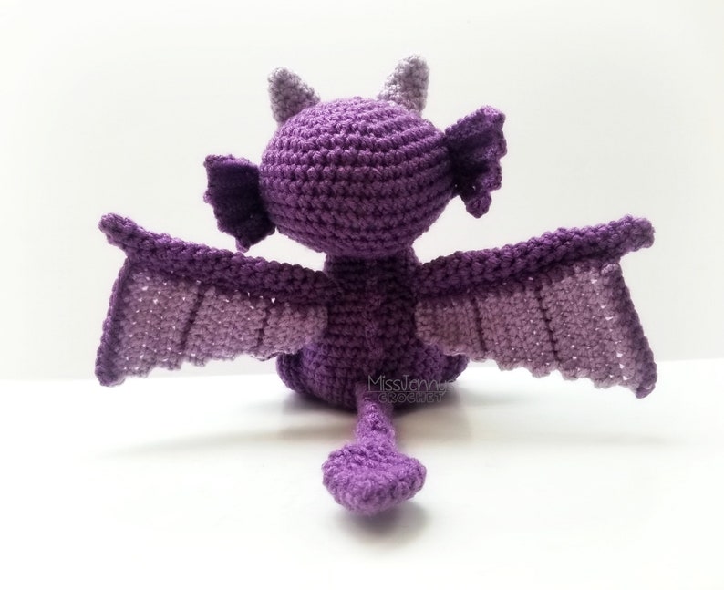 May include: A handmade crochet dragon in shades of purple, viewed from the back. The dragon has outstretched wings, small horns, and a tail. The wings and body are a gradient of purple hues.