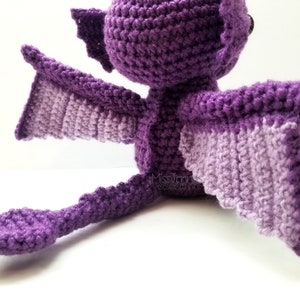 May include: A handmade crochet dragon in shades of purple. The dragon has large wings with a lighter purple underside, a long tail, and small horns. The dragon is sitting down, showcasing its detailed crochet work.