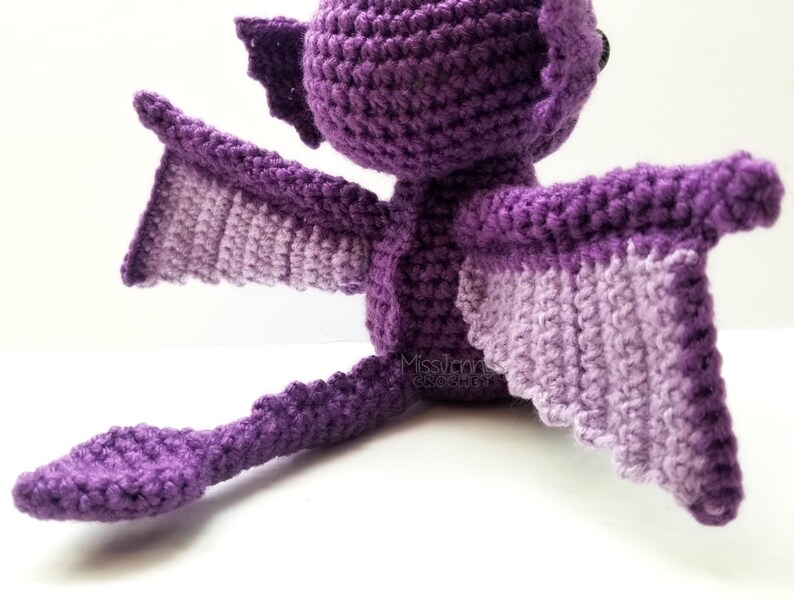 May include: A handmade crochet dragon in shades of purple. The dragon has large wings with a lighter purple underside, a long tail, and small horns. The dragon is sitting down, showcasing its detailed crochet work.