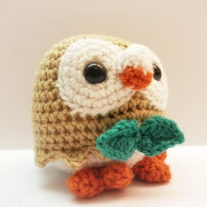 May include: A handmade crochet owl in tan, white, orange, and green. The owl has large black eyes, white facial features, and orange feet. A green bow is attached to the owl's chest. The owl is a cute, whimsical, and decorative item.