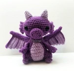 May include: A handmade crochet dragon in shades of purple. The dragon has large black eyes, small horns, and outstretched wings. The body is a lighter shade of purple, and the wings are a darker shade. The dragon is a whimsical, handcrafted item.