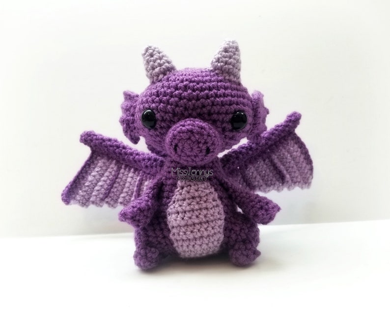 May include: A handmade crochet dragon in shades of purple. The dragon has large black eyes, small horns, and outstretched wings. The body is a lighter shade of purple, and the wings are a darker shade. The dragon is a whimsical, handcrafted item.